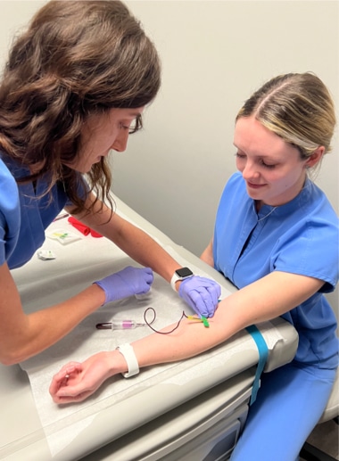 A healthcare student in blue scrubs and purple gloves is drawing blood from a woman also in blue scrubs.