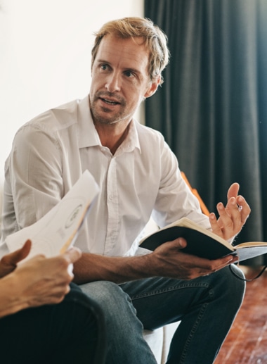 A man with light brown hair and a beard, wearing a white shirt and jeans, is sitting and holding an open book, gesturing with his other hand.