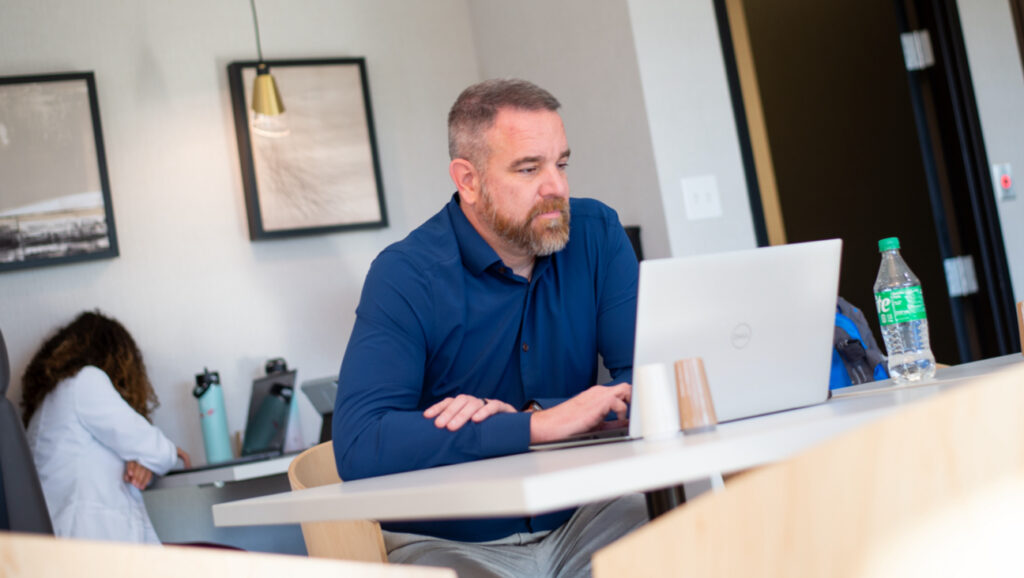A man with a beard, wearing a blue shirt, sits at a desk working on a silver laptop.