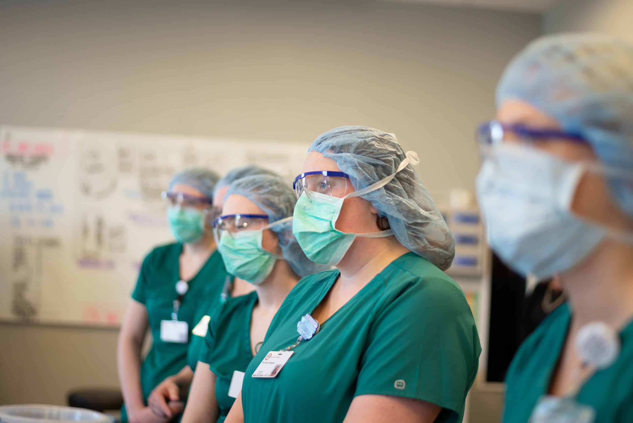 A group of students wearing green scrubs, surgical caps, masks, and protective goggles stand in a row, attentively listening or watching something. A whiteboard with writing is visible in the background.