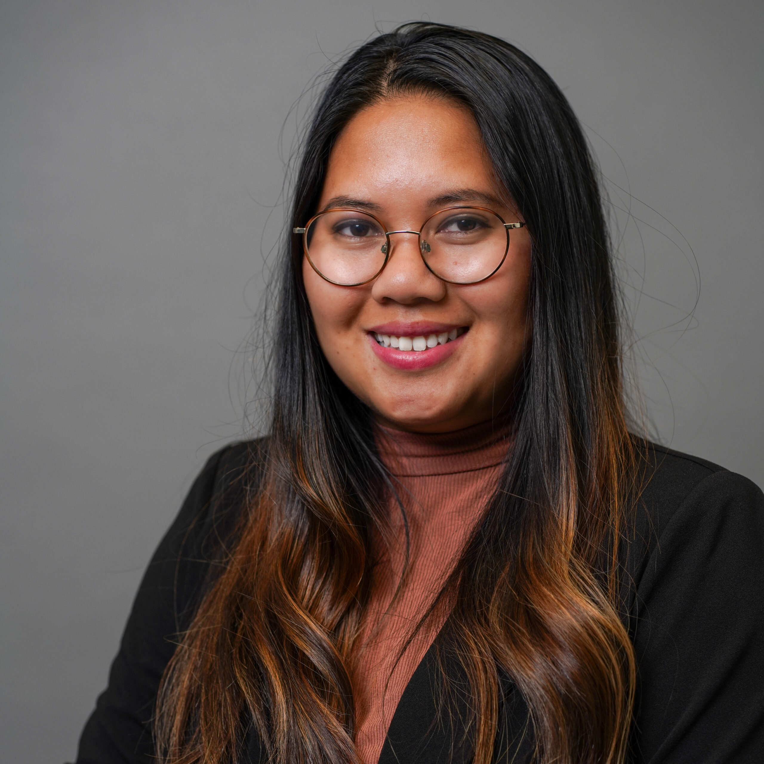 A person with long, dark hair and glasses is smiling. They are wearing a brown turtleneck and a black blazer. The background is plain gray.