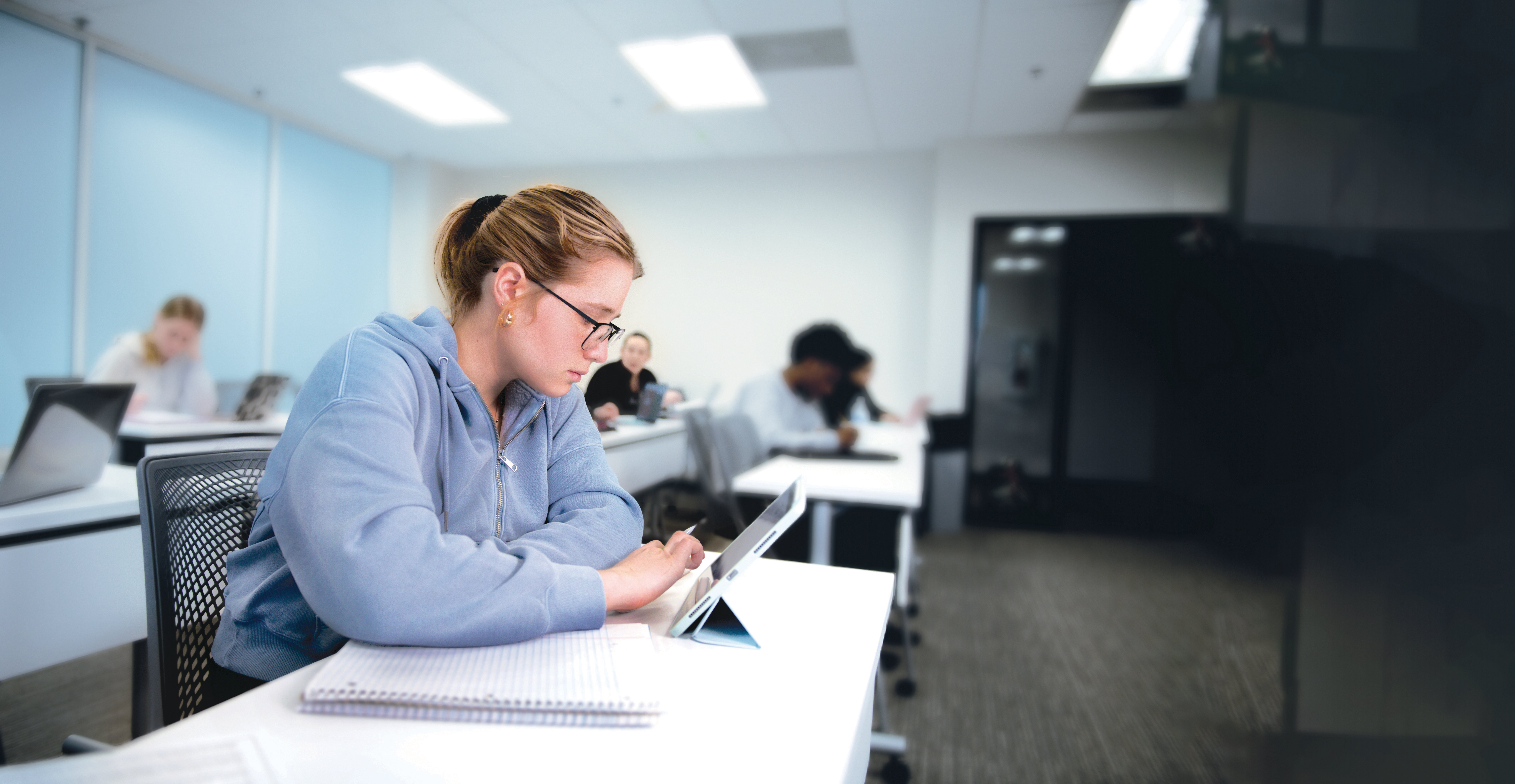 A student wearing glasses and a blue hoodie sits at a desk in a classroom, focused on a tablet. Open notebooks and a pencil are on the desk. Other students sit and work at their desks in the background. The classroom has white walls and bright lighting.