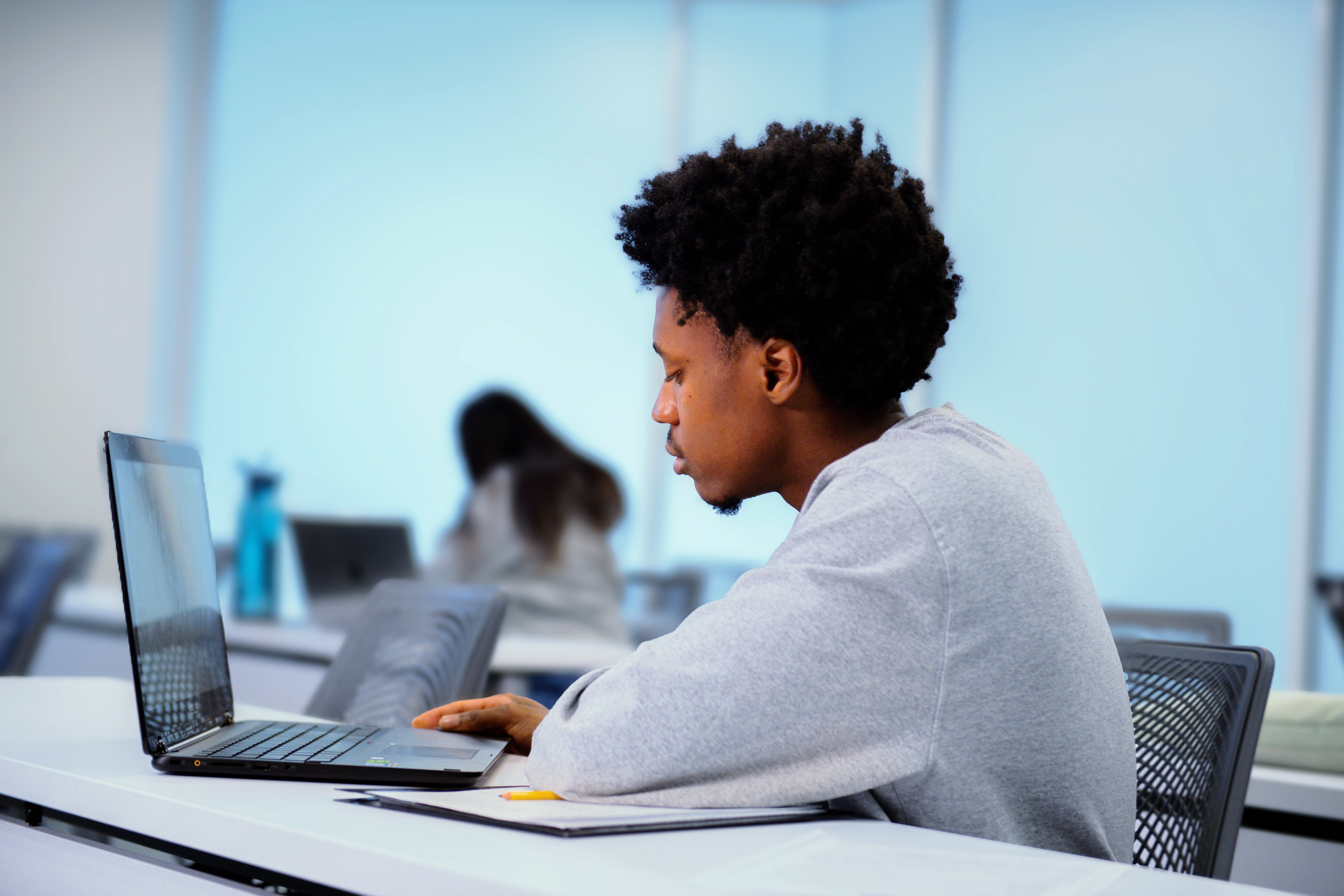 A student with short curly hair sits in an empty classroom, focused on a laptop. They are wearing a casual long-sleeved shirt. In the background, a blurred figure is visible, suggesting another individual working in the same room.
