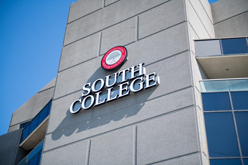 An exterior view of a tall grey building with reflective windows. The building bears a logo reading "South College" in large, white letters below a red circular emblem. The sky is clear and blue.