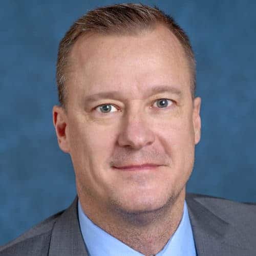 Portrait of a man with short brown hair and blue eyes, wearing a grey suit jacket and light blue dress shirt. He is looking directly at the camera with a neutral expression. The background is a solid dark blue color.