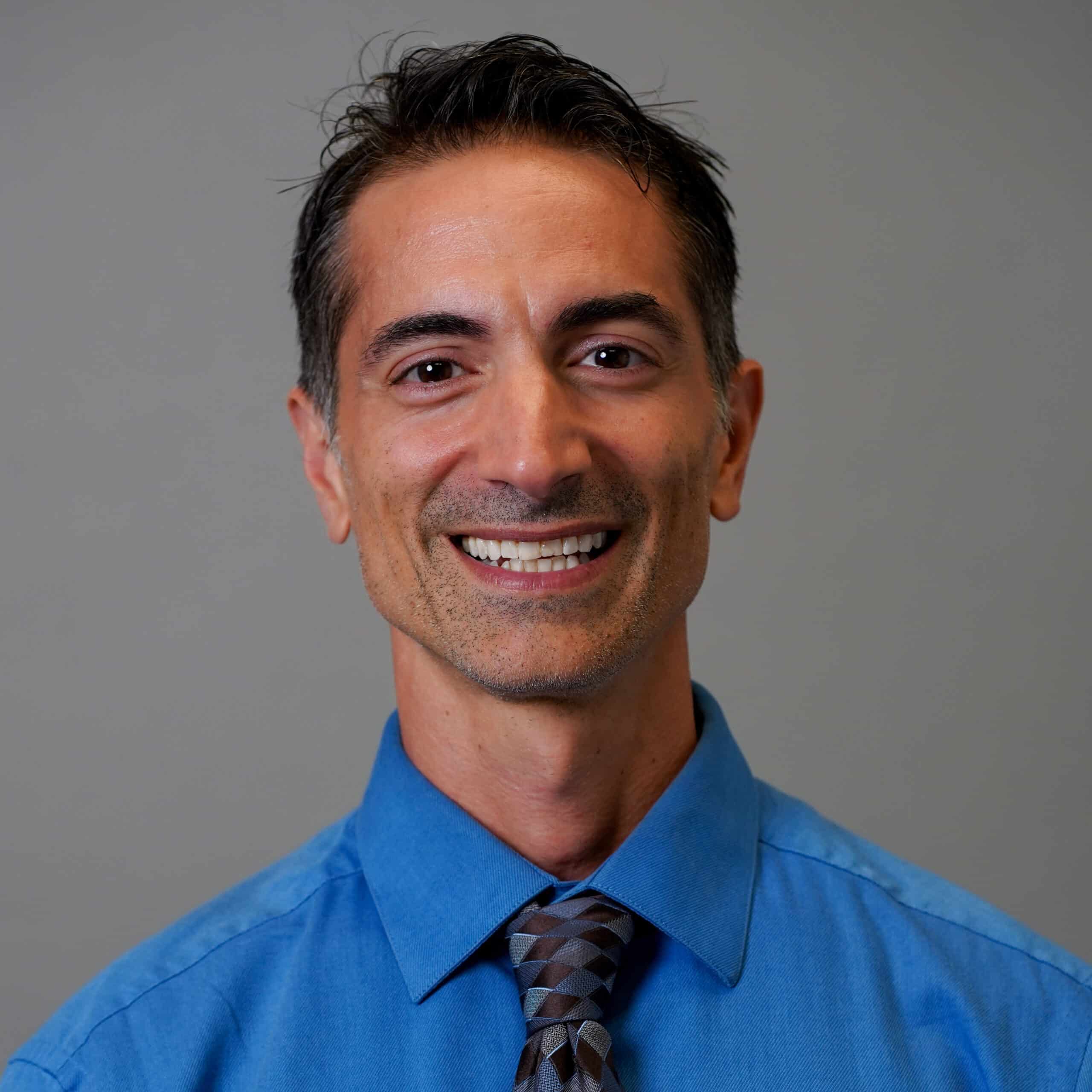 A man with short dark hair and a graying beard, smiling at the camera. He is wearing a bright blue dress shirt and a dark patterned tie. The background is a plain gray color.