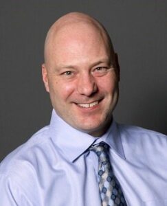 Cheryl Sparks A man with a friendly smile is wearing a light blue dress shirt and a patterned tie. He is posed against a neutral gray background.