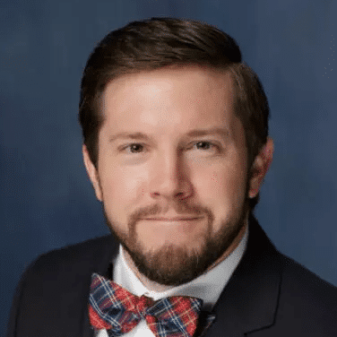 A man with short brown hair and a beard is wearing a navy blue blazer, a white shirt, and a red and blue plaid bow tie. He is smiling slightly and posing against a plain blue background.