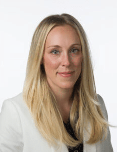 A woman with long, straight blonde hair, wearing a white blazer and a dark blouse underneath, is looking straight into the camera with a neutral expression. The background is plain white.