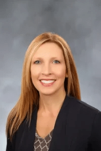 A woman with long blonde hair is smiling at the camera. She is wearing a dark blazer over a patterned blouse. The background is a neutral gray color.