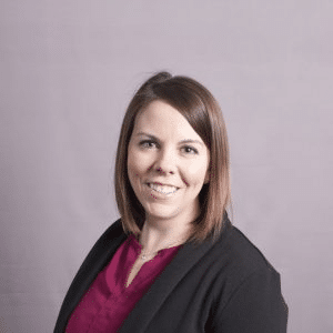A woman with shoulder-length brown hair is smiling at the camera. She is wearing a maroon top and a black jacket. The background is plain and light-colored.