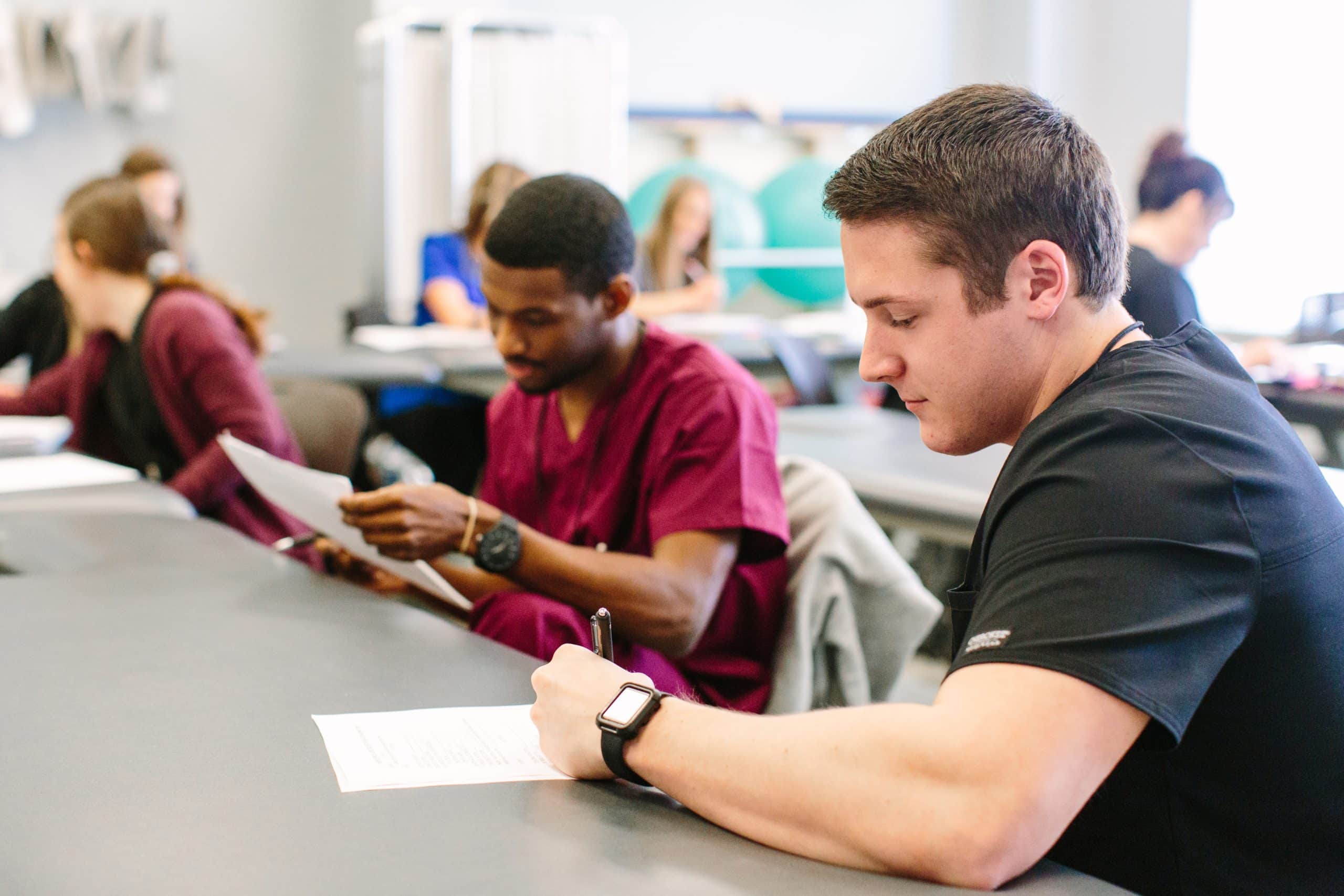 A group of people are seated at a table in a classroom setting. The focus is on two individuals. One person is in black scrubs and writing on a paper, while the other is in maroon scrubs, reading a document. Other attendees are visible in the background.