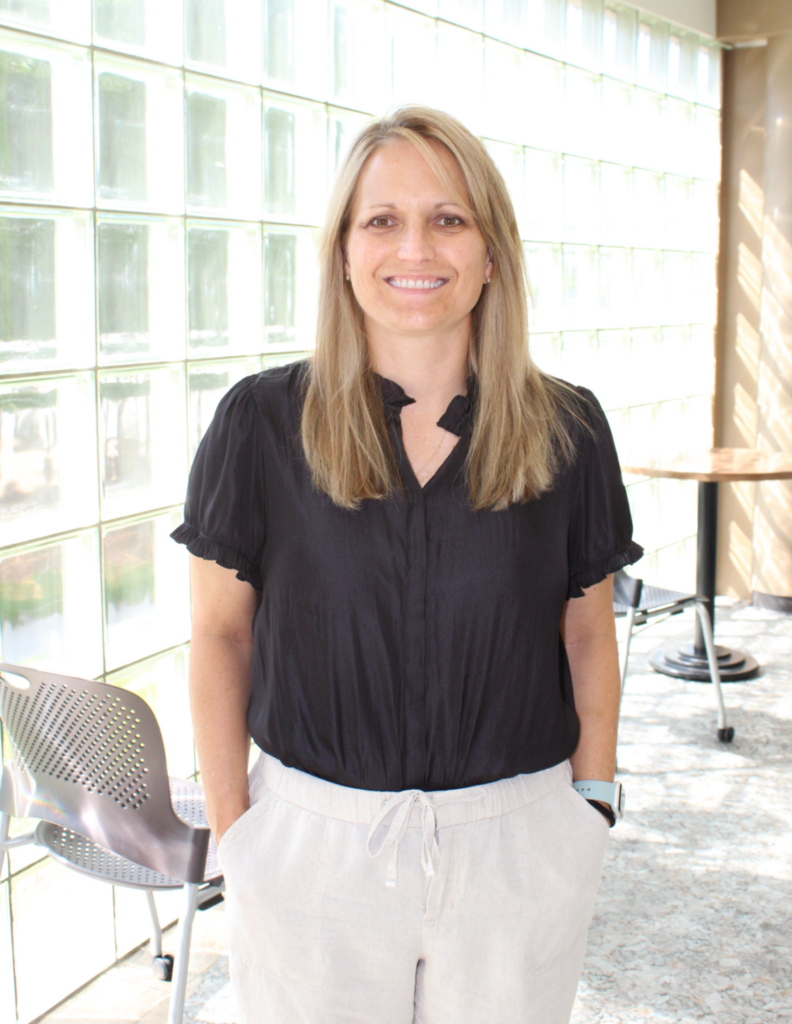 A smiling woman with shoulder-length blonde hair is wearing a colorful blouse with patterns in pink, black, and white. The blouse has cut-out shoulders.