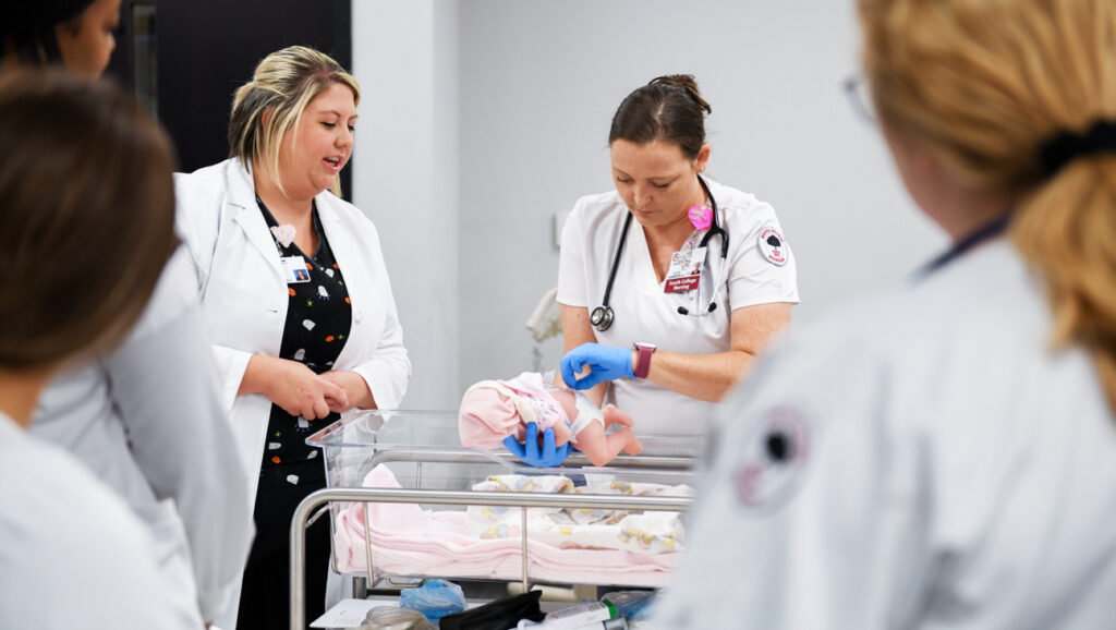 A group of Nursing students observe a nurse while she tends to an infant mannequin in a medical training setting.