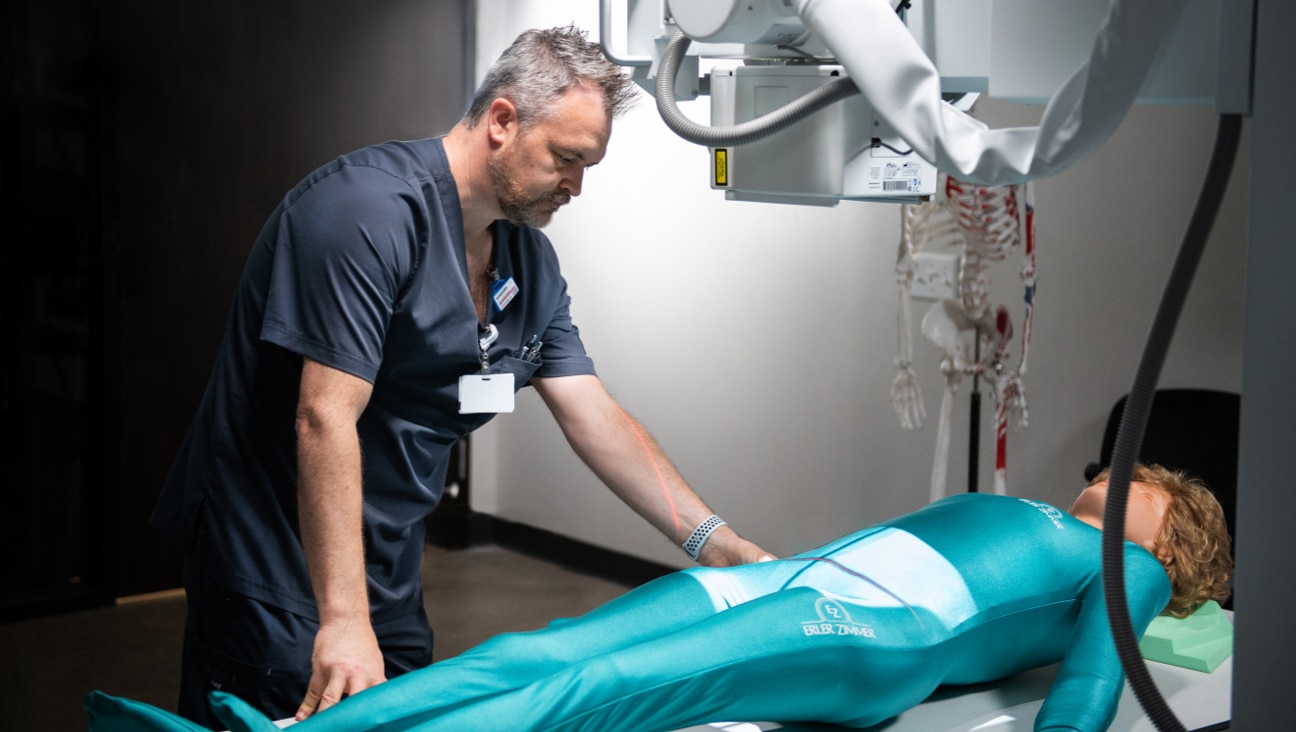 A Radiography student in dark scrubs examines a patient on an examination table.