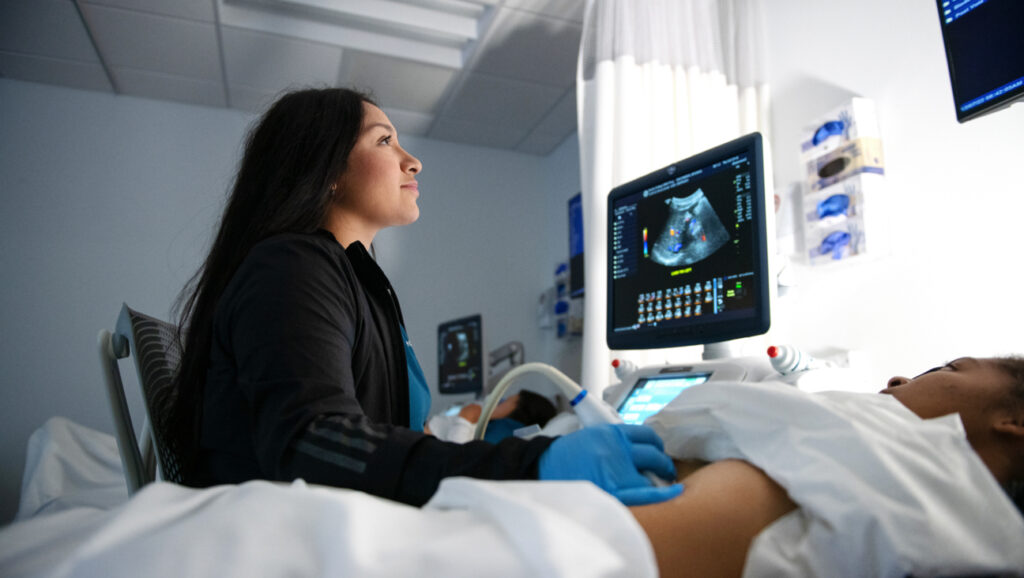 An Imaging Sciences student uses an ultrasound machine to perform a sonogram on a patient's abdomen in a medical examination room.