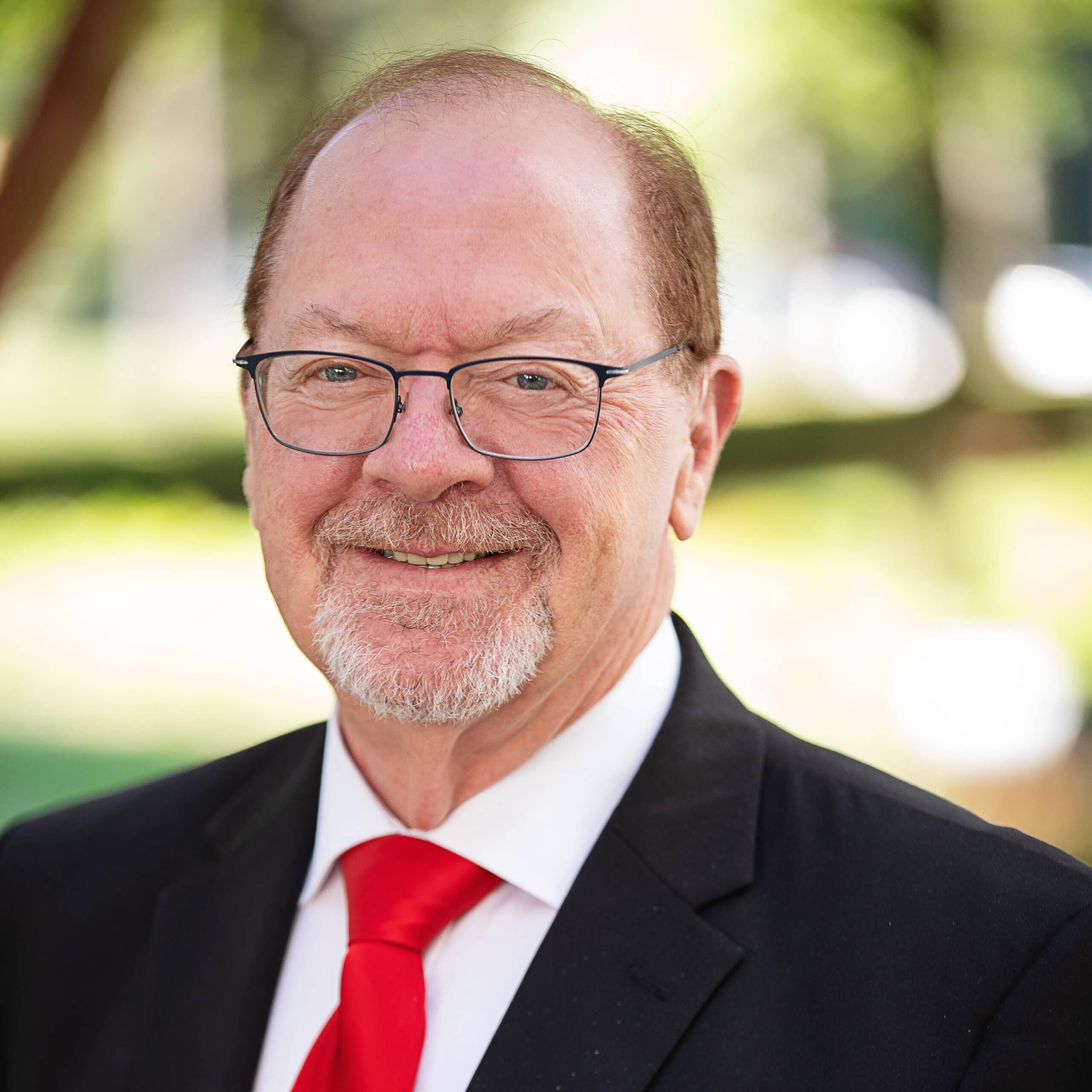 A smiling man with glasses, a short beard, and thinning hair is wearing a black suit jacket, white shirt, and a bright red tie. The background is a blurred outdoor setting with greenery and sunlight.