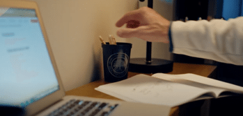 A person reaches for a pencil from a cup on a desk. An open laptop and a notebook with sketches are nearby, while a lamp provides light. The scene suggests a work or study environment.