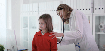 A doctor is using a stethoscope to listen to a young girl's back in a brightly lit medical office. The girl is wearing a red sweater and has shoulder-length hair. Shelves with binders and medical equipment are visible in the background.