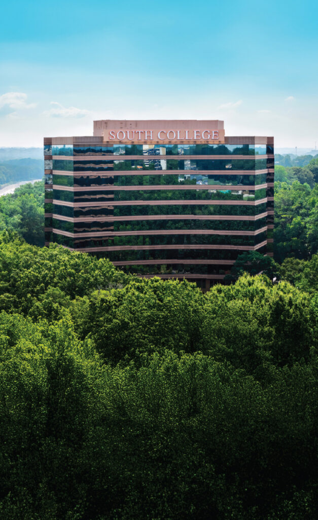 A glass office building with “South College” on top, surrounded by dense green trees under a bright blue sky.