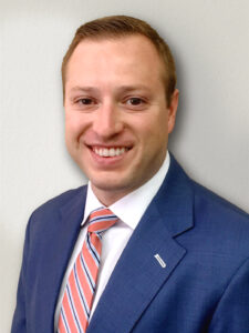 A smiling man in a blue suit and a striped tie stands against a plain light background.