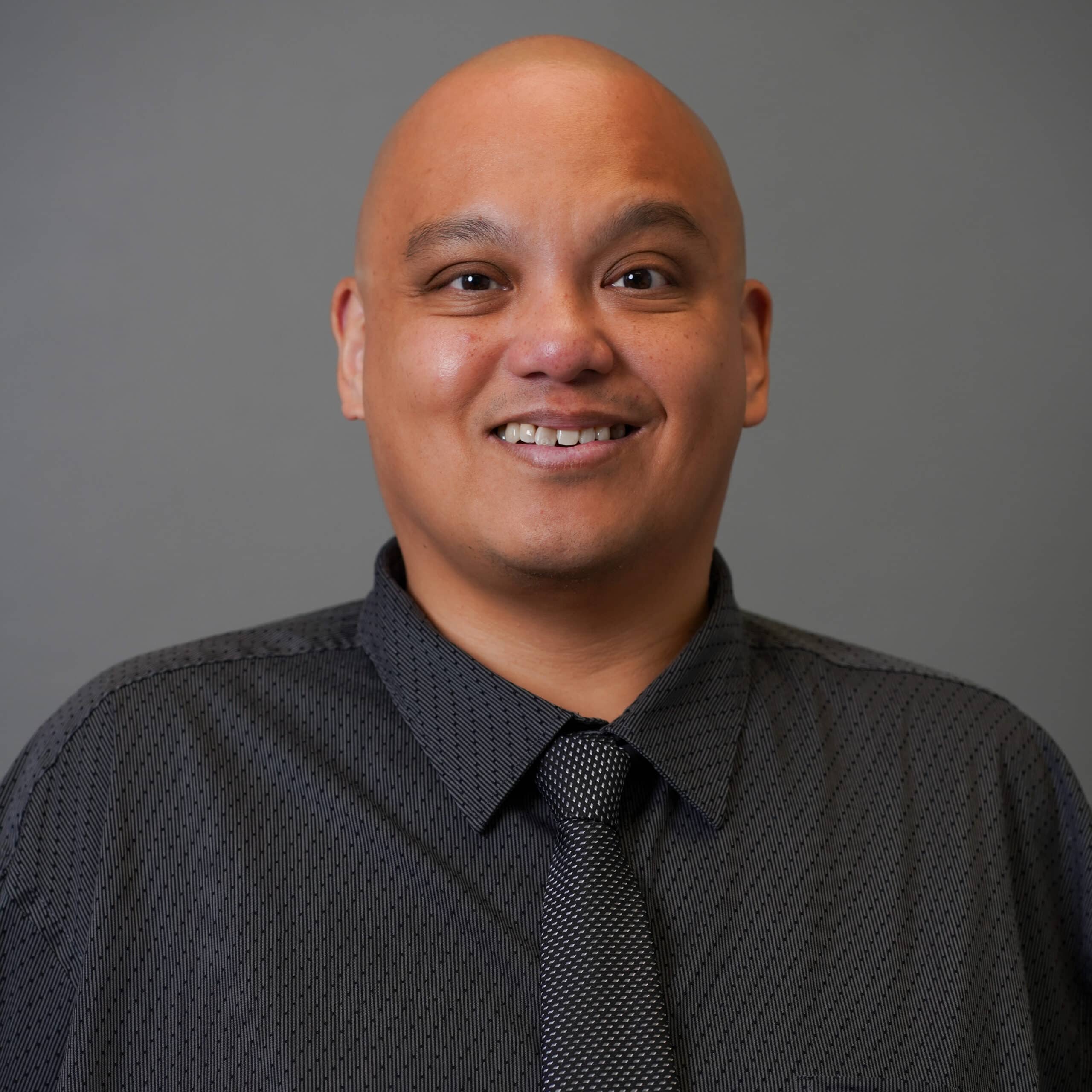 A person with a friendly smile is wearing a dark patterned shirt and a matching tie. The background is a plain gray color.