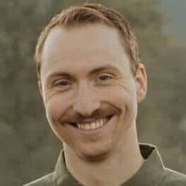 A person with short, red hair and a mustache is smiling. They are wearing a green button-up shirt with a collar. The background appears to be outdoors with greenery.
