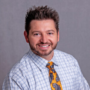 Miranda, Nicole A man with short, spiky hair and a trimmed beard smiles at the camera. He is wearing a light blue checkered dress shirt and an orange tie with blue paisley patterns. The background is plain and gray.
