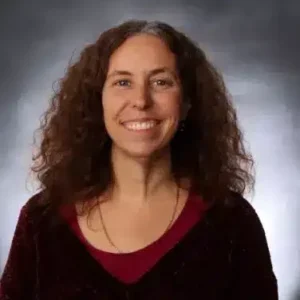 A woman with long, curly hair and a broad smile is wearing a red top layered with a darker, velvety sweater. The background is a neutral, shaded gradient.