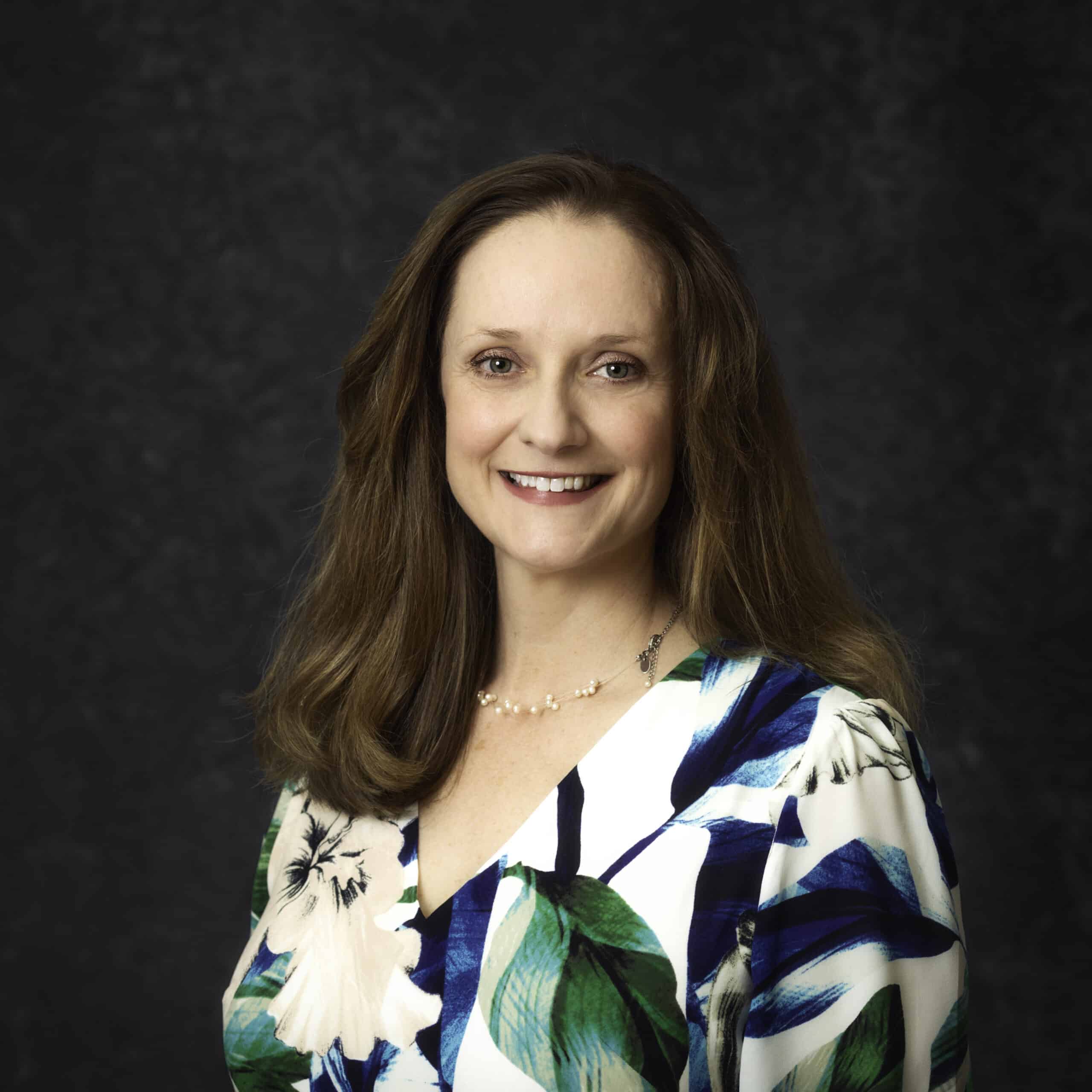 A person with long brown hair is smiling against a dark background. They are wearing a floral blouse with shades of blue and green.