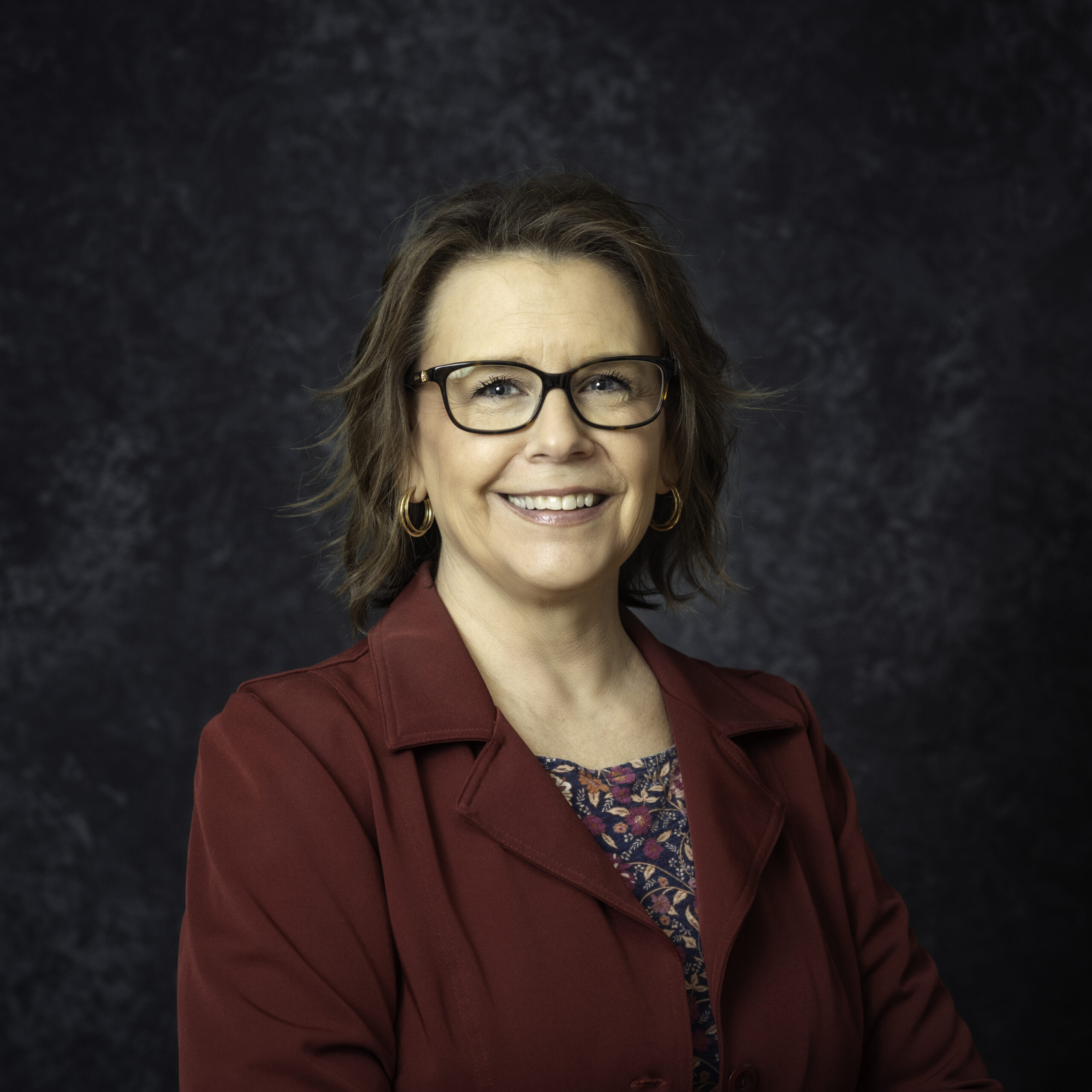 A woman with glasses and shoulder-length hair smiles while wearing a burgundy jacket over a floral top.