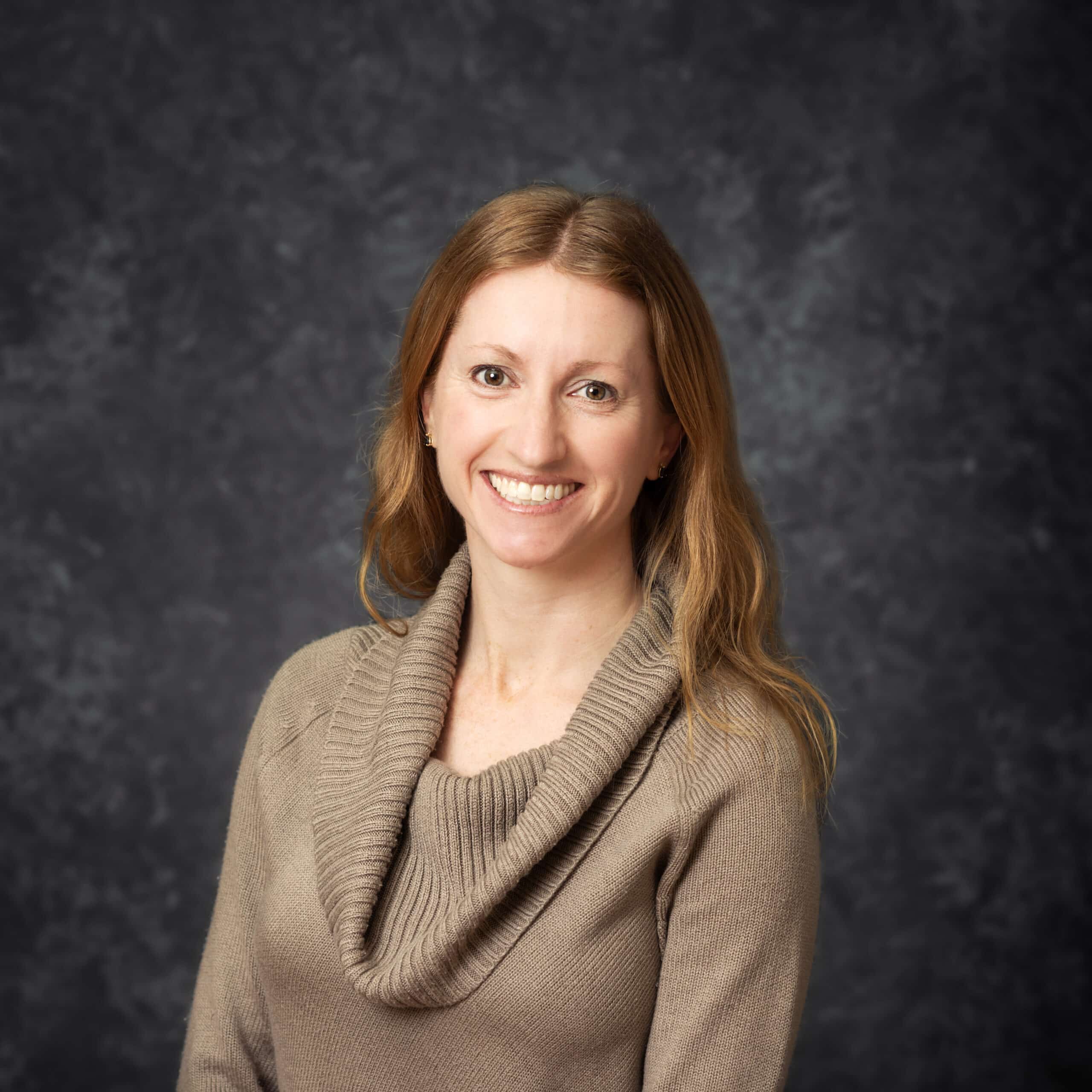 A woman with long light brown hair smiles while wearing a tan sweater.