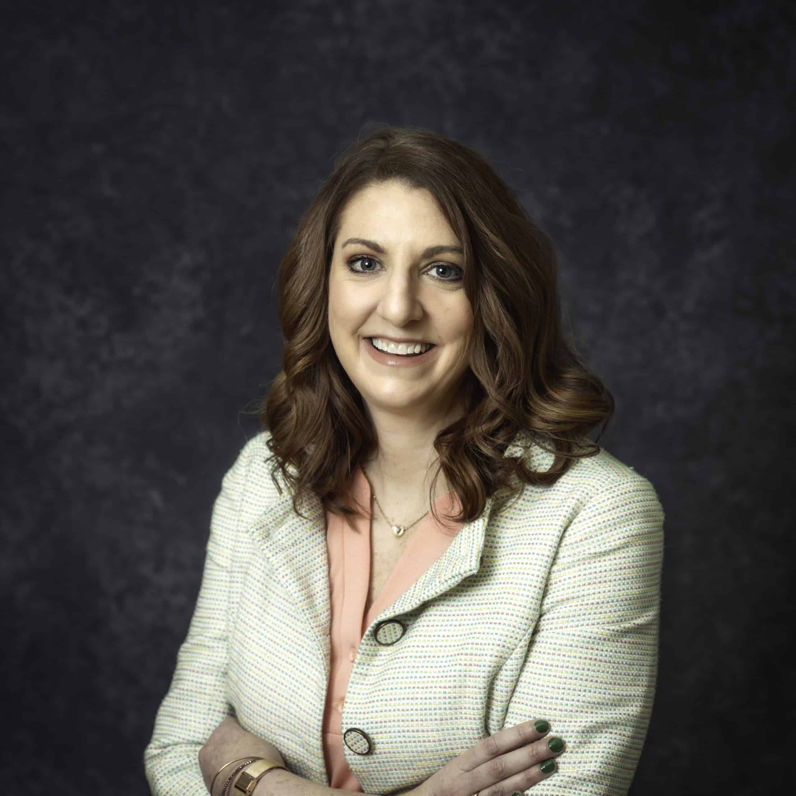 A person with shoulder-length wavy hair, wearing a light-colored jacket over a peach top, is smiling at the camera with arms crossed. The background is dark and textured.