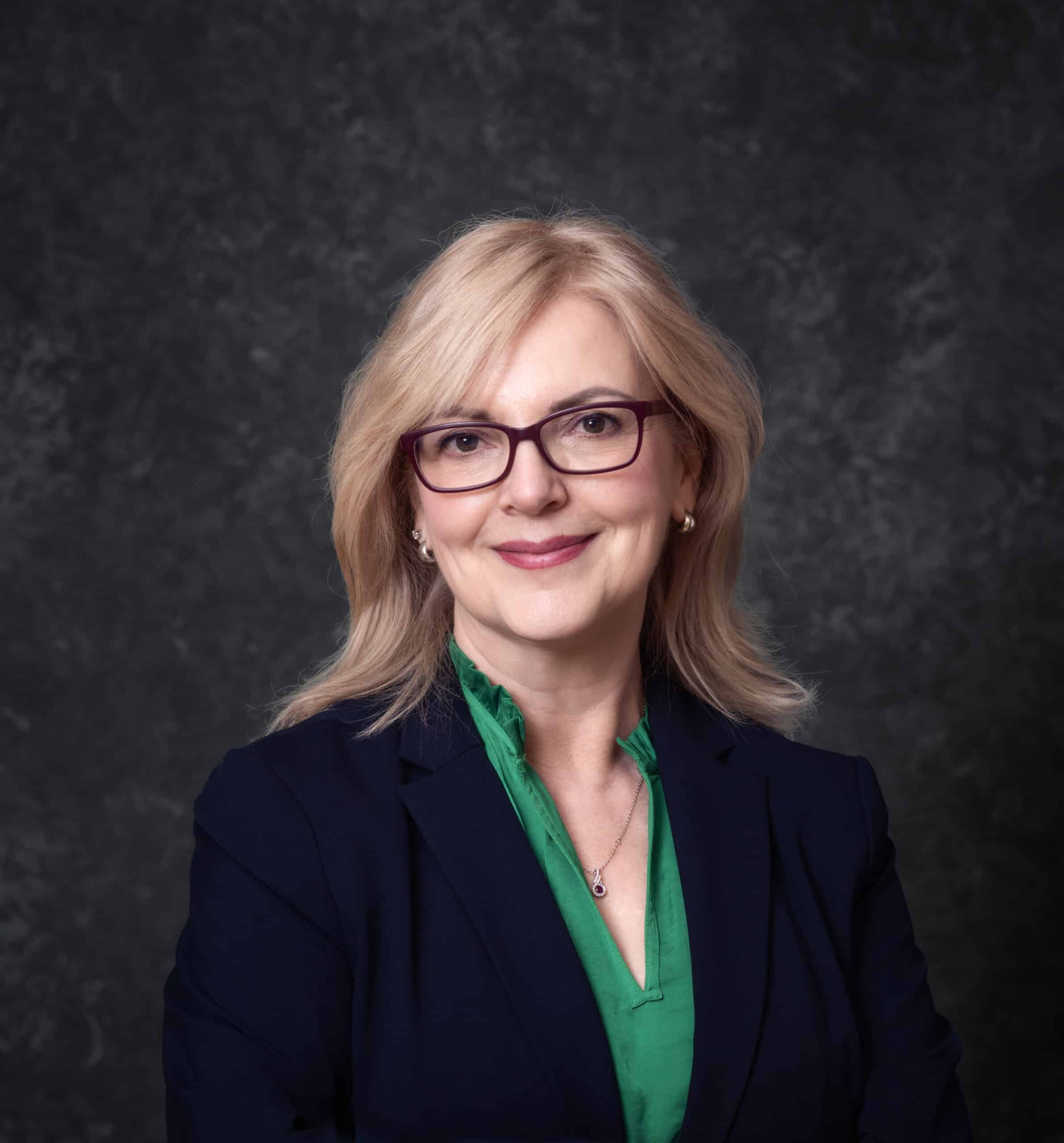 A woman with shoulder-length blonde hair, wearing glasses and a navy blazer over a green blouse, smiles softly against a dark textured background.