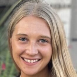 A young woman with long, straight blonde hair smiles at the camera. She has blue eyes and is outdoors, with greenery and blurred background visible behind her.