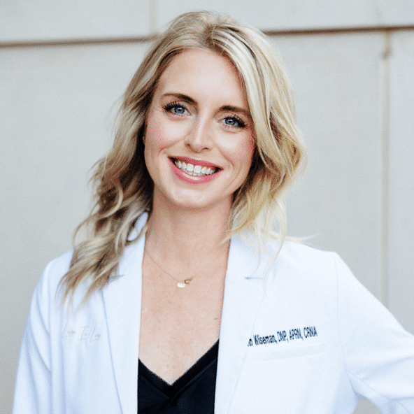 A person smiling at the camera, wearing a white lab coat over a black top. The person has medium-length blonde hair and is standing against a plain, light-colored background.