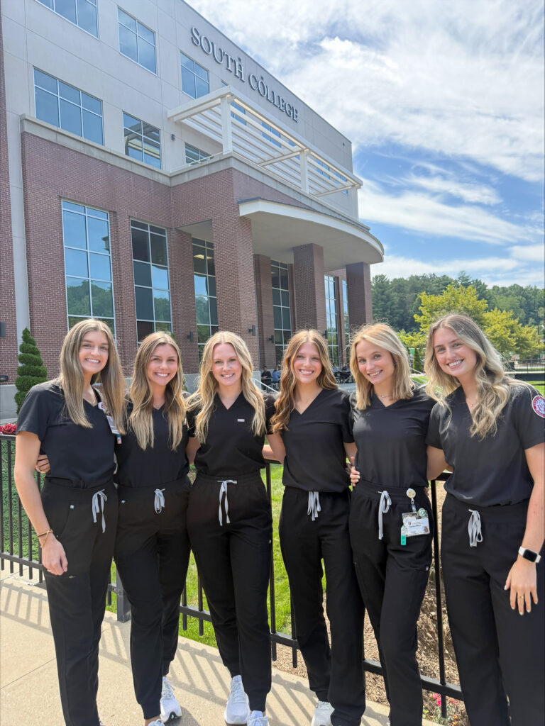 Six women in matching black scrubs stand smiling together outside a building labeled “South College” on a sunny day, with greenery and blue sky in the background.