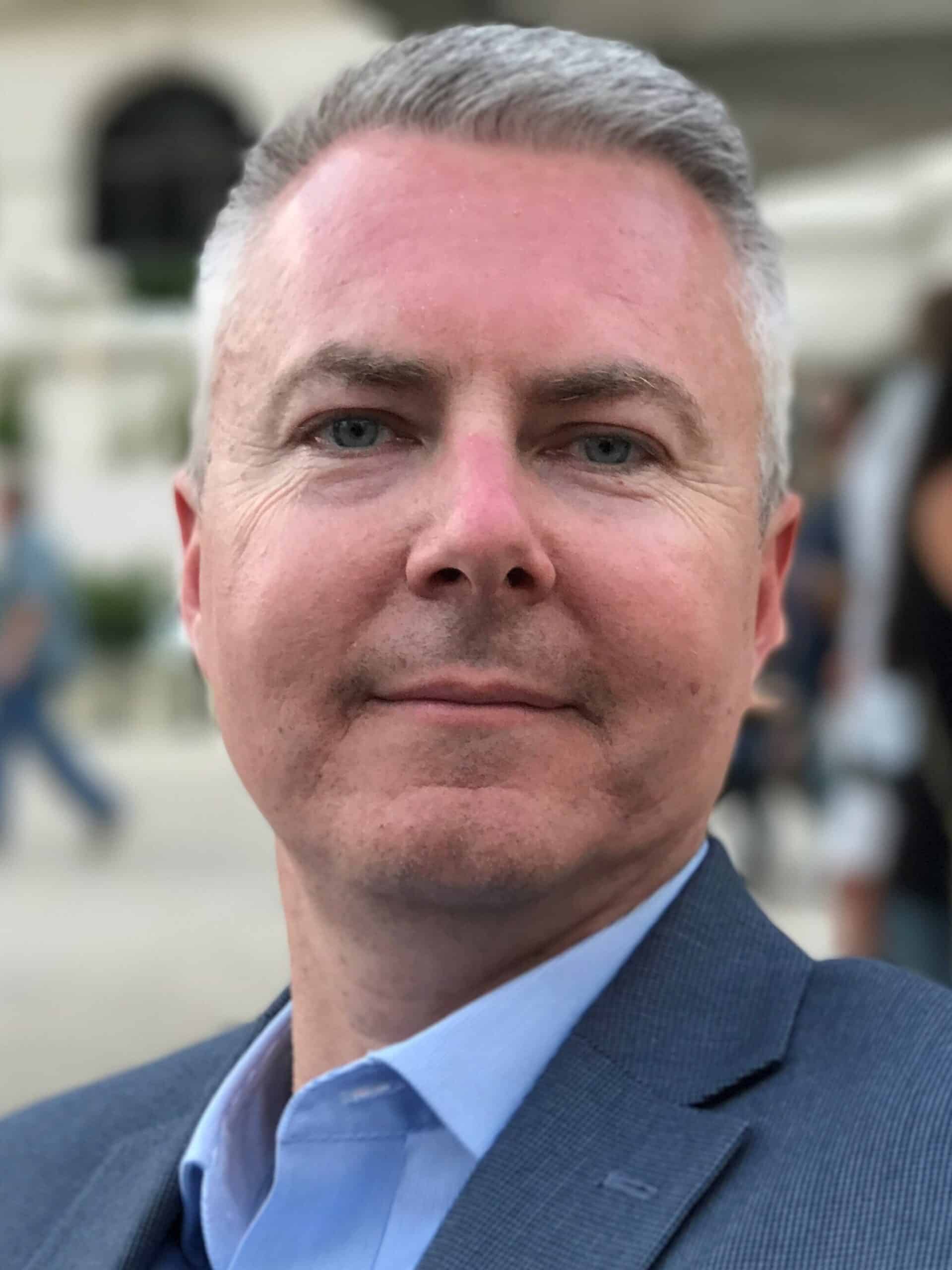 A close-up of a man with short gray hair and a trimmed beard, wearing a blue dress shirt and gray blazer. He is looking directly at the camera with a slight smile. The background is blurred with people and a building with arches in soft focus.