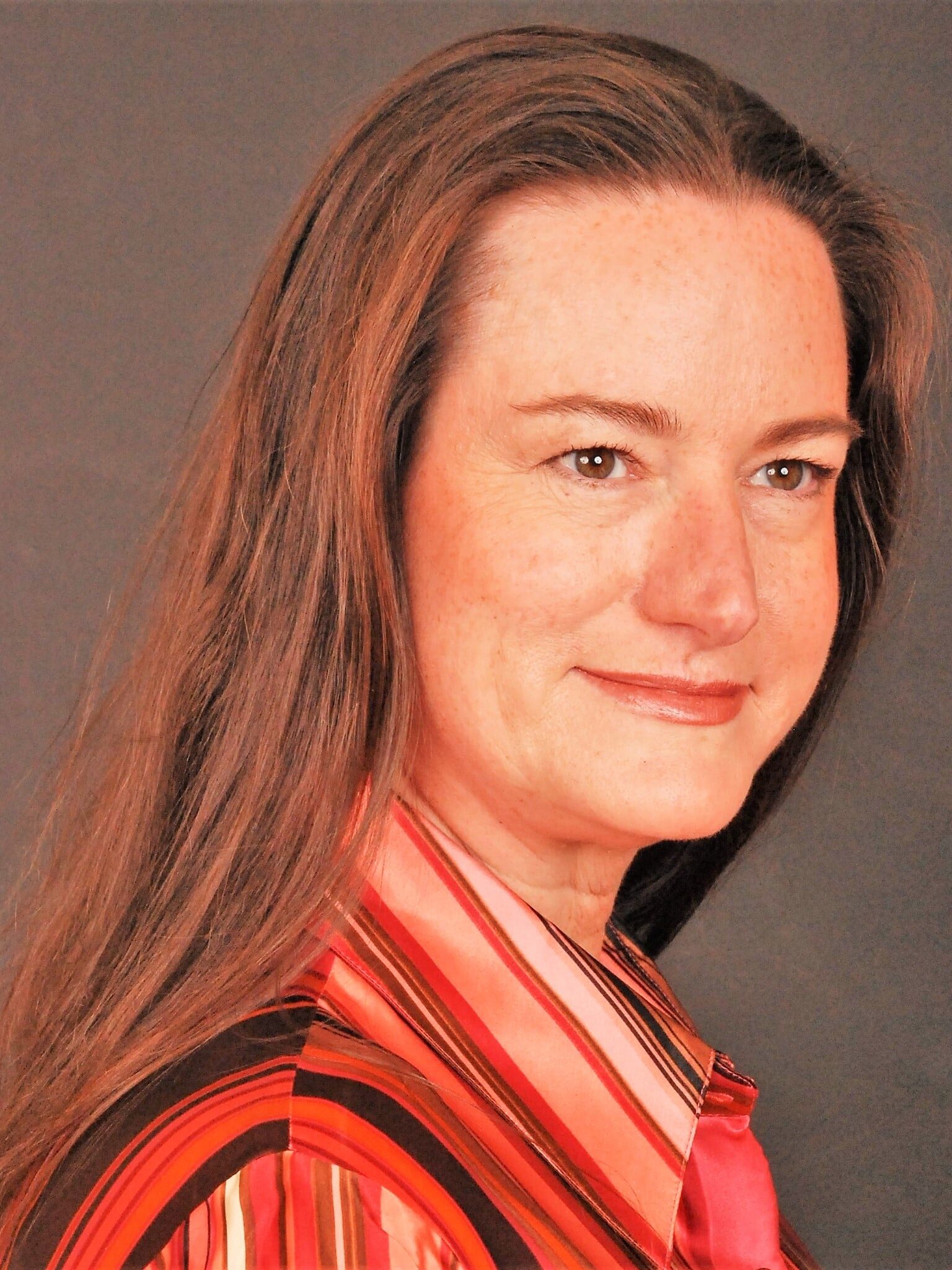A woman with long, straight brown hair and fair skin is smiling slightly. She is wearing a red and pink striped shirt. The background is a solid gray color.