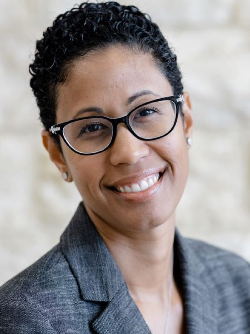 A person with short curly hair and black-rimmed glasses is smiling at the camera. They are wearing a grey blazer and a white shirt, with a light-colored background behind them.