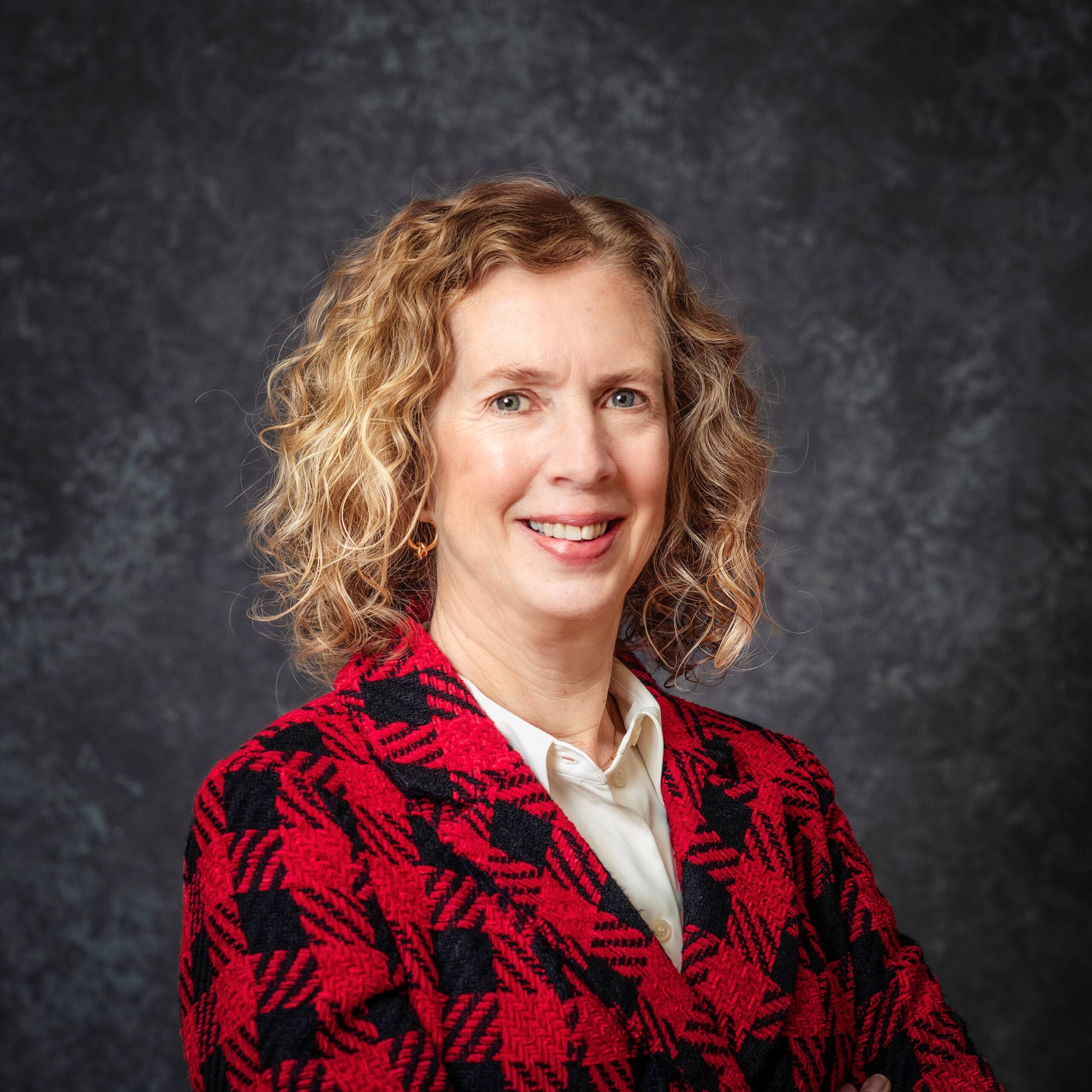 A woman with curly blonde hair smiles at the camera. She is wearing a red and black plaid jacket over a white blouse, standing against a dark, textured background.