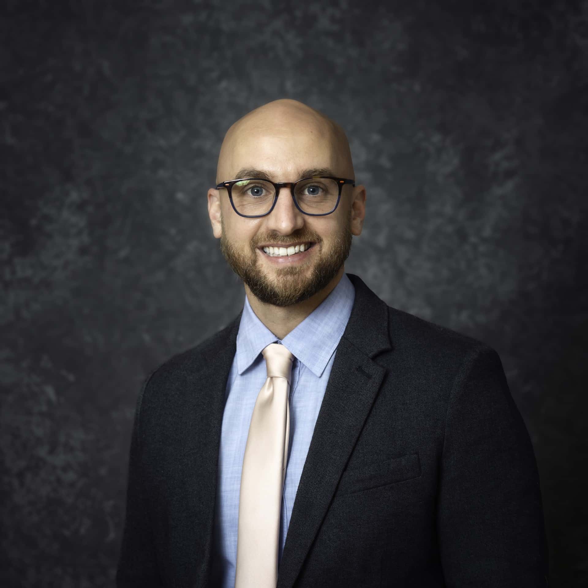 A smiling man with glasses and a trimmed beard is wearing a dark suit, light blue shirt, and a pale tie, posed against a dark, textured background.