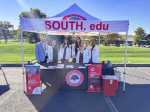 A group of people, mostly in white coats, stand smiling behind a South College booth under a branded tent outdoors. The table displays brochures and banners promoting South College programs.