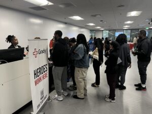 A group of people stands in line at an indoor job fair, gathered near a Grady Health System booth with a sign reading "HEROES WORK HERE." Other booths and tables are visible in the background.
