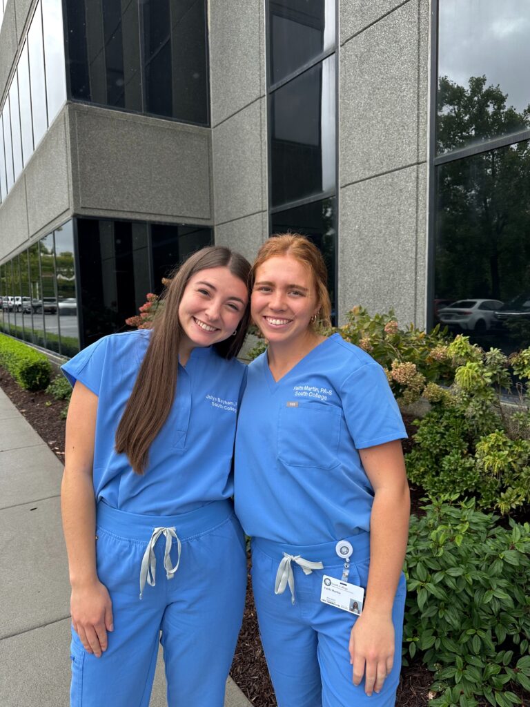 Two women wearing blue scrubs stand smiling together outside a modern building, surrounded by green bushes and flowers. One has a badge clipped to her pocket, and both appear happy and relaxed.