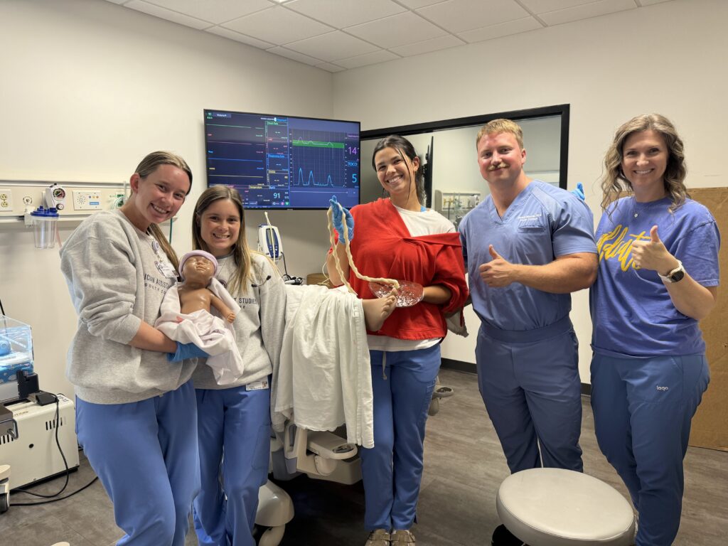Five smiling medical professionals in scrubs pose in a clinic room, holding medical models including a baby and pelvis. Monitors, medical equipment, and a towel are visible in the background. One person gives a thumbs up.
