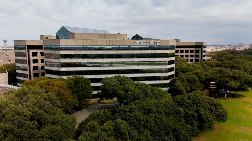 A large, modern building labeled "South College" sits behind green trees under a cloudy sky. The building has reflective windows and appears to be part of a campus surrounded by greenery.