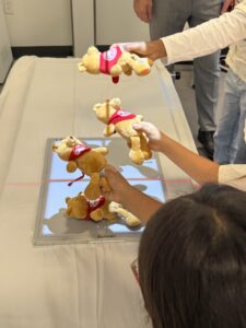 Several people hold up identical teddy bears with red shirts above a flat scanning device, which is placed on a white cloth-covered surface. One person’s head and arm are visible in the foreground.