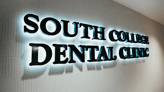 Illuminated black letters on a patterned wall read “South College Dental Clinic.”.