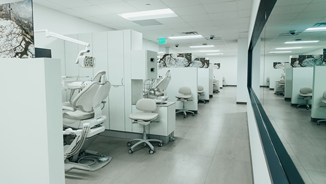 A modern dental clinic with white walls, dental chairs, equipment, and partitions. The area is clean, bright, and organized, with large windows reflecting the room.